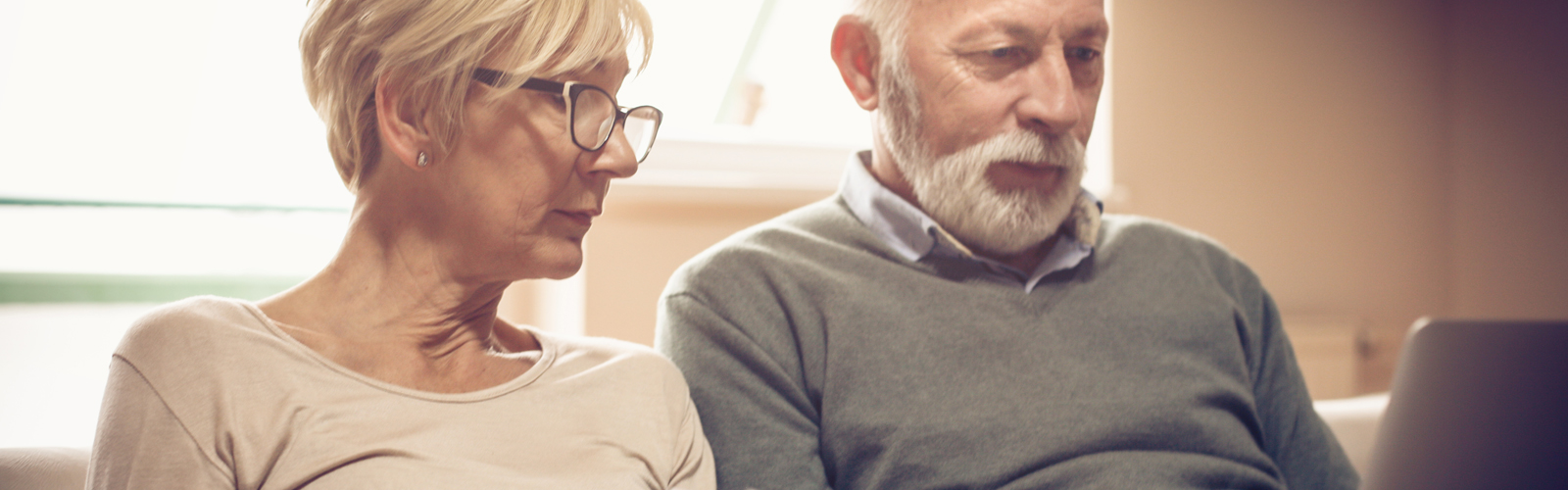 An older couple, sat next to each other on a sofa, look at some paperwork