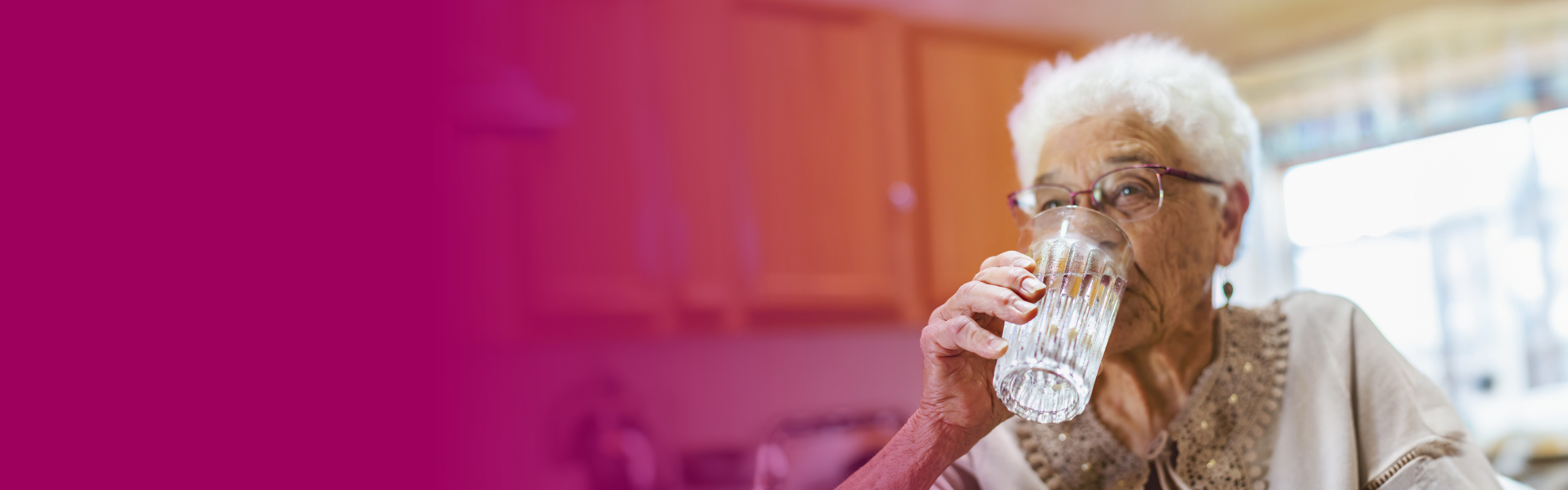 An older woman sat in a kitchen, drinking a glass of water