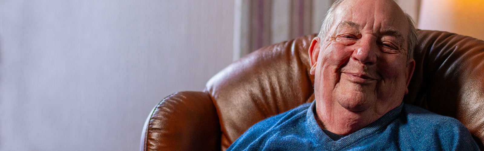 An older man in a blue shirt, sat in an armchair, smiling