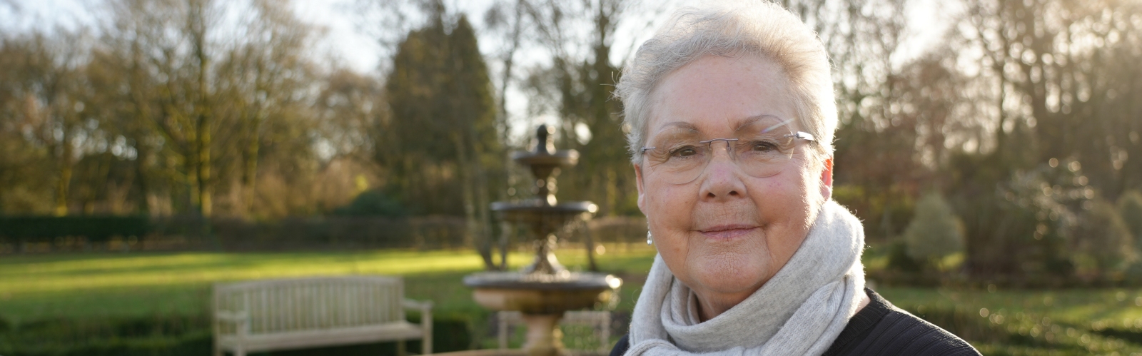 An older lady with short hair and glasses stands in a park, looking to camera