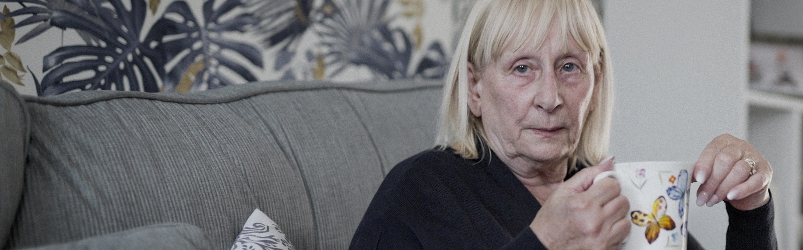 An older woman with light blonde hair holds a cup of tea, looking sadly into the distance