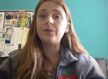 A lady with red hair sits in front of a green wall with a noticeboard on it