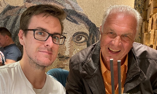 A young man with glasses smiles with his grandfather, enjoying a meal out