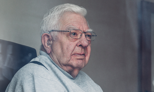 An older man sits waiting alone in his living room, staring out of his back door